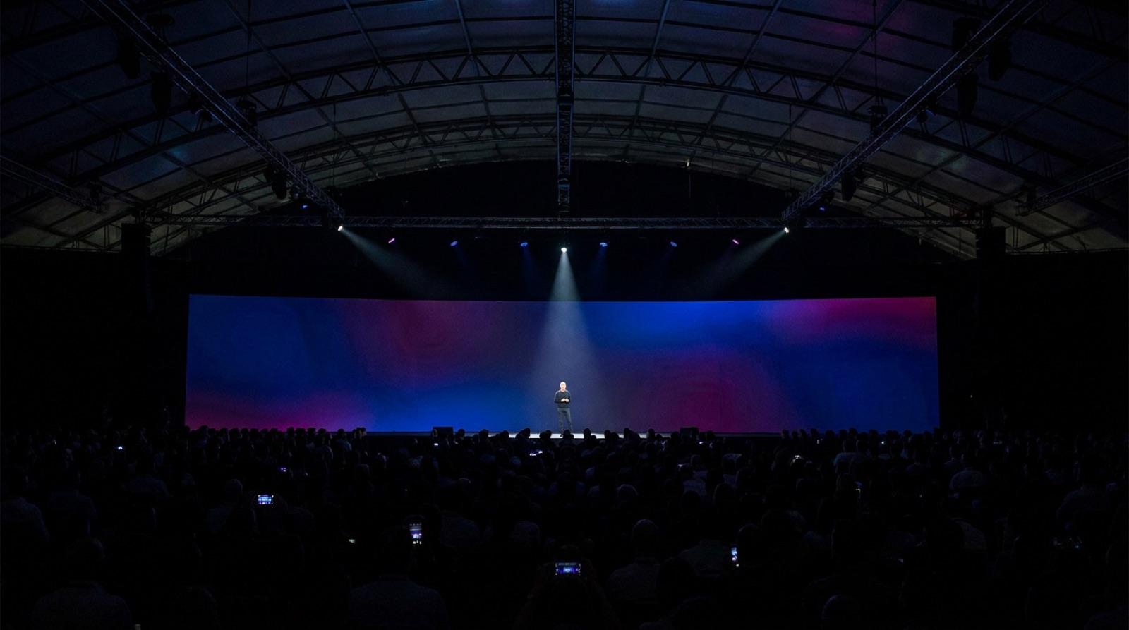 A keynote speaker on a wide stage, a single spotlight, the audience a dark sea of faces